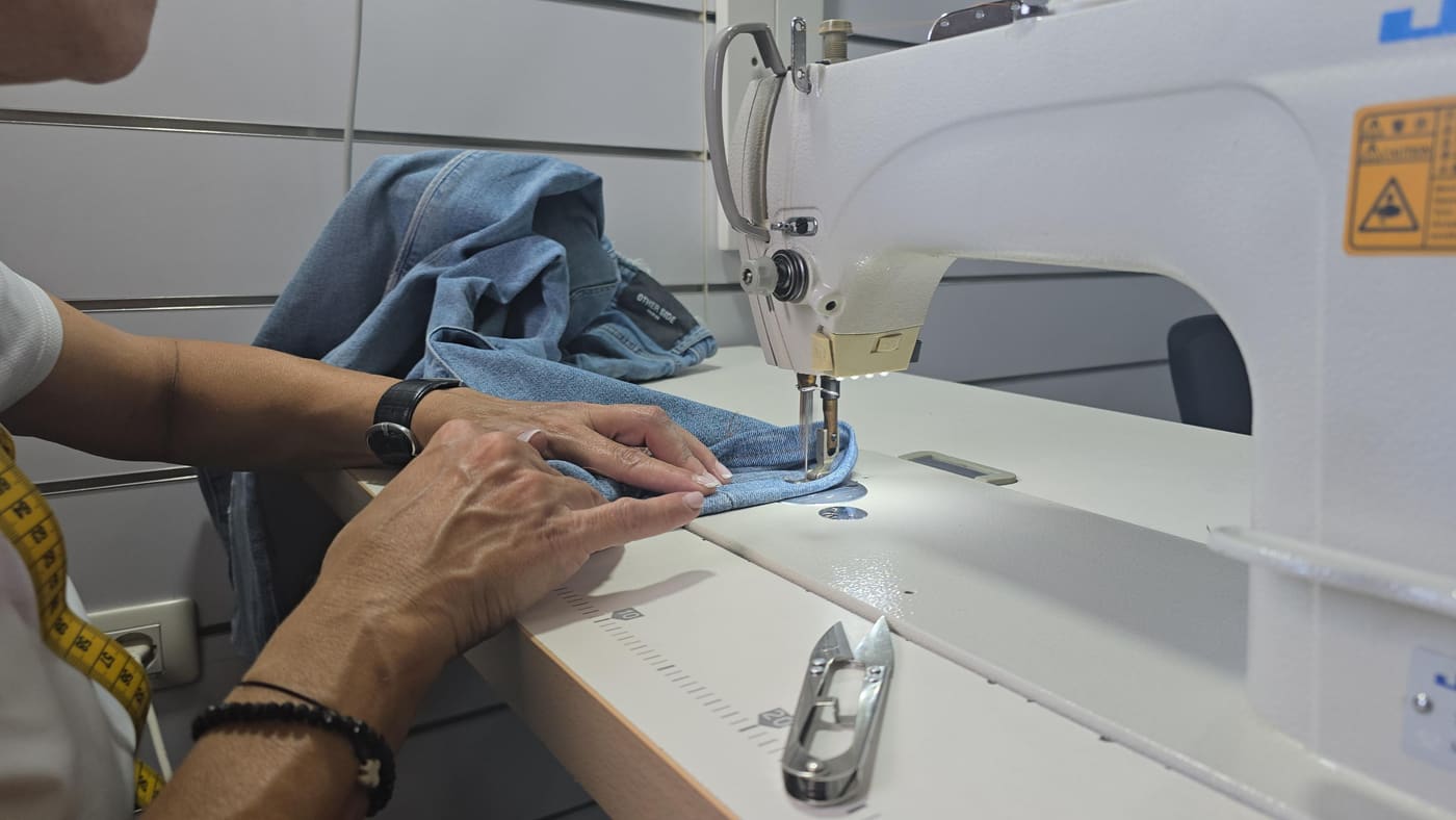 Hands feeding a denim shirt through a sewing machine for alterations, a tape measure to the side